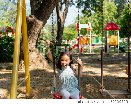 Cute girl having fun on the swing in the playground in the park. Children playing in the playground during summer vacation. Cute girl having fun on the swing in the playground in the park. Children playing in the playground during summer vacation. 120139733