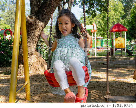 Cute girl having fun on the swing in the playground in the park. Children playing in the playground during summer vacation. Cute girl having fun on the swing in the playground in the park. Children playing in the playground during summer vacation. 120139734