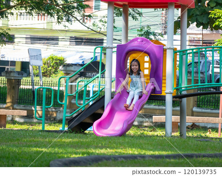 Cute girl having fun on a colorful slide in an outdoor park. Children playing in the playground during summer vacation. Cute girl having fun on a colorful slide in an outdoor park. Children playing in the playground during summer vacation. 120139735