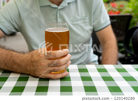 Man holding a glass of beer, sitting at a table with a green checkered tablecloth, outdoors at a cafe 120140280