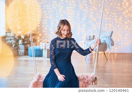 teenage girl in a dress on a swing in front of a Christmas tree and presents surrounded by golden Christmas lights teenage girl in a dress on a swing in front of a Christmas tree and presents surrounded by golden Christmas lights 120140461