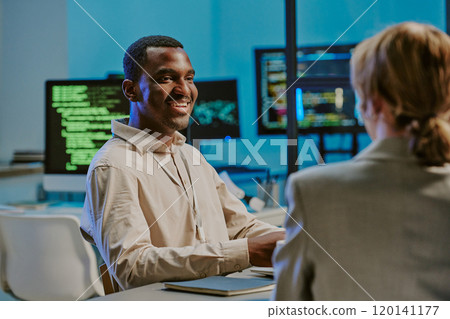 Cheerful young African American male cybersecurity specialist smiling while having conversation with colleagues during meeting 120141177