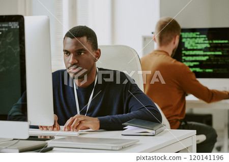 Young African American male IT specialist typing code on desktop computer at workplace in office 120141196