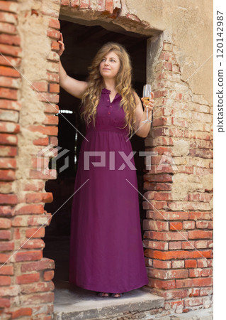 Elegant woman in purple dress holding champagne in rustic brick archway.Wine Day. 120142987