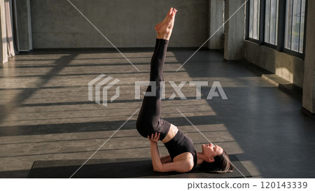 Pilates exercise. Physical practice. Athletic woman in activewear perform shoulder stand gymnastic pose laying on yoga mat in gym fitness studio indoors. Pilates exercise. Physical practice. Athletic woman in activewear perform shoulder stand gymnastic pose laying on yoga mat in gym fitness studio indoors. 120143339