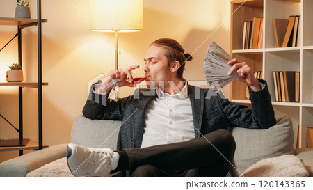 Success relax. Good result. Handsome young man in modern suit holding money enjoying red wine in glass sitting on sofa in office. 120143365