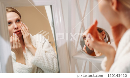 Eye patches. Face care. Moisturizing treatment. Woman clean healthy fresh skin applies collagen hydrogel pads mask in white soft robe looking in mirror. 120143410