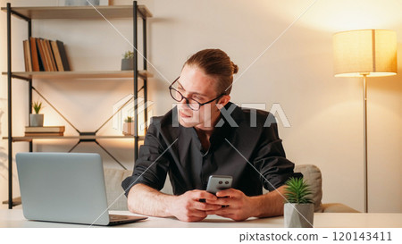 Business work. Technology story. Successful young handsome man in glasses in black shirt holding smartphone while looking at laptop sitting in office. 120143411