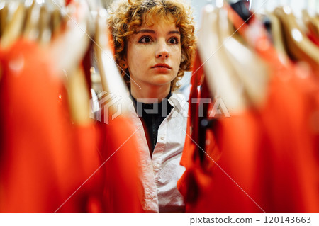 young attractive red-haired woman choosing red clothes in boutique young attractive red-haired woman choosing red clothes in boutique 120143663