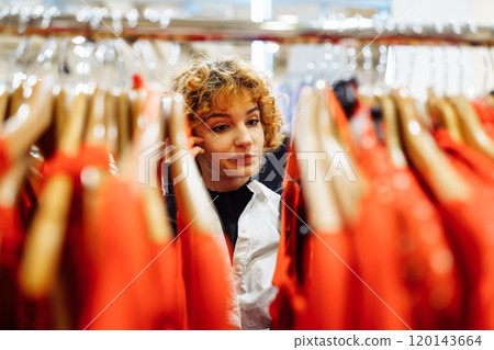 young attractive red-haired woman choosing red clothes in boutique young attractive red-haired woman choosing red clothes in boutique 120143664