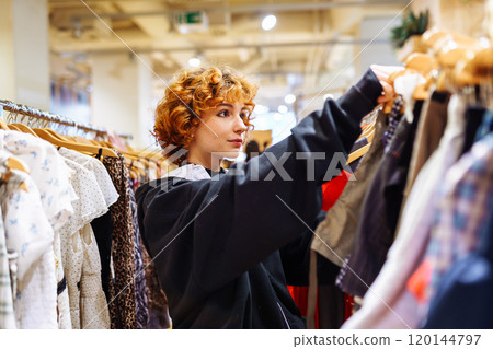 portrait of red-haired attractive young woman trying on clothes in store 120144797