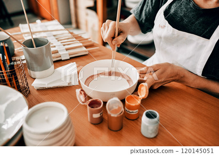 I use art to escape. Cropped shot of an unrecognizable woman sitting alone and painting a pottery bowl in her workshop. 120145051