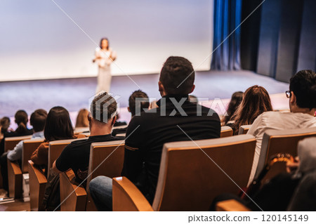 Business and entrepreneurship symposium. Female speaker giving a talk at business meeting. Audience in conference hall. Rear view of unrecognized participant in audience. 120145149