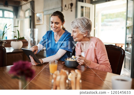 Its a great way to stay in touch with family. Shot of a young nurse and senior woman using a digital tablet at breakfast time in a nursing home. 120145633