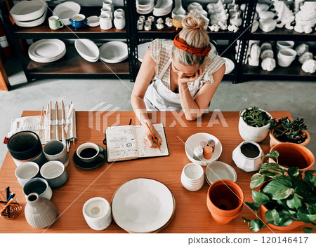 The hard work never ends. High angle shot of an unrecognizable woman sitting and sketching in a notebook in her pottery workshop. 120146417