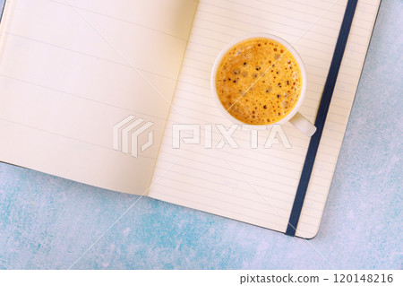 Cup of steaming coffee espresso sits next to an open notebook planner on light blue surface 120148216