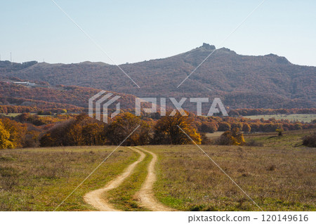 Winding dirt path through vibrant autumn landscape with distant mountains under clear sky 120149616