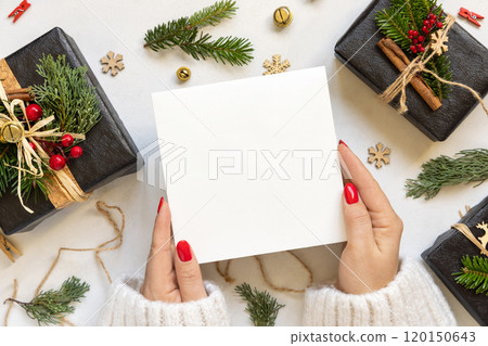 Hands with square card near gift boxes, fir branches and Christmas decor, Mockup 120150643