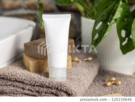 White cream tube on brown towels near vessel sink and green plant close up, cosmetic mockup 120150648