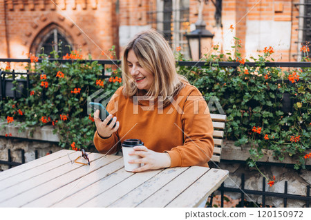 Woman holding bio coffee paper cup and smartphone, she is sitting on the cafe terrace on the city street. Urban concept. Happy Girl sitting at a table with a coffee using mobile phone. 120150972