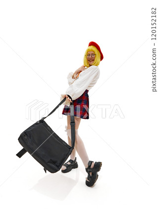 Woman in quirky schoolgirl outfit with yellow wig, red beret and plaid skirt posing playfully with bag against white studio background. Woman in quirky schoolgirl outfit with yellow wig, red beret and plaid skirt posing playfully with bag against white studio background. 120152182