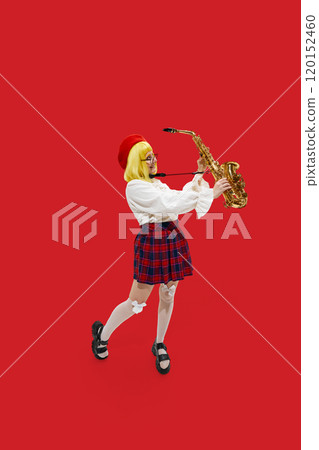 Happy, excited woman dressed stylish vibrant outfit with yellow wig posing with saxophone against vibrant red background. 120152460