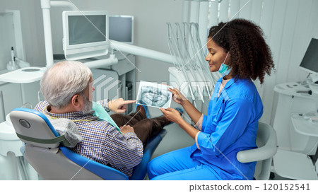 Dentist reviews X-ray image with mature patient, explaining details and preparing them for next steps in their dental care. Dentist reviews X-ray image with mature patient, explaining details and preparing them for next steps in their dental care. 120152541