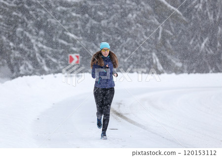 Woman Jogging Along a Snowy Road During a Blizzard in Winter Mountains 120153112