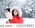 Beautiful brunette in winter in forest in red jacket with hood on her head with smartphone in hand. 120154091