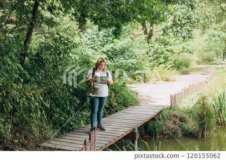Stylish hipster woman holding paper map and phone, wearing backpack and relaxing in nature. Travel and wanderlust concept. Amazing chill moment, summer time. Full body 120155062