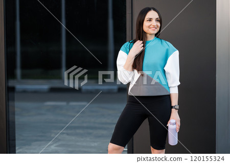 Woman holding bottle with water after morning workout. Young athletic woman standing near dark wall background after jogging Healthy lifestyle concept 120155324