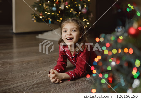 Happy girl of 7 years old in red pyjamas lies on floor of house next to Christmas tree with side of garlands at home Happy girl of 7 years old in red pyjamas lies on floor of house next to Christmas tree with side of garlands at home 120156150