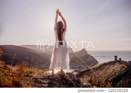 A woman stands on a hill overlooking the ocean. She is wearing a white dress and has her hands on her head. The scene is serene and peaceful, with the woman looking out over the water. 120156329