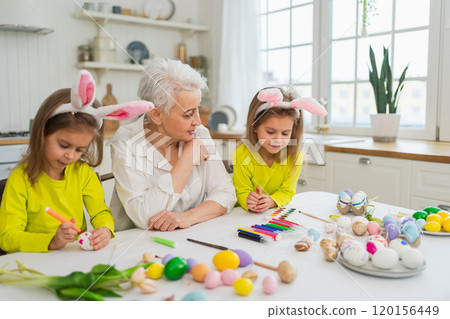 Happy Easter. Two little girls sisters twins grandmother painting eggs enjoying time together. Happy family grandma granddaughters child kids preparing for Easter. Spring Christian festival tradition 120156449