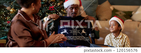 Header Image of African American man trying on Christmas sweater while celebrating Christmas with family at home and exchanging gifts 120156576