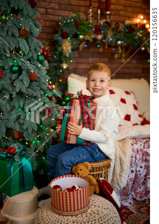 Cute boy hugging a gift box next to Christmas tree. A child in anticipation of miracles and a 120156635