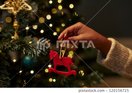 Close up of childs hand holding festive Christmas ornament of red toy horse while decorating Christmas tree with twinkling lights in background copy space 120156725