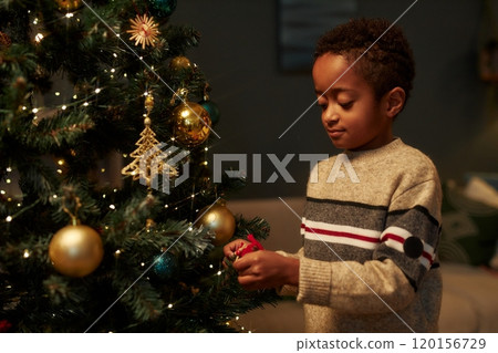 Waist up portrait of cute African American boy decorating Christmas tree in cozy home setting with dim lighting copy space 120156729