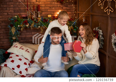 Happy family opening presents next to Christmas tree and smiling cheerfully on Christmas Eve in a 120156739