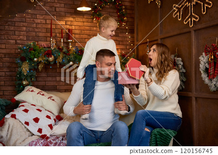 Happy family opening presents next to Christmas tree and smiling cheerfully on Christmas Eve in a 120156745