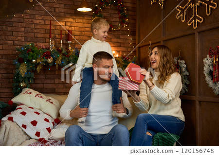 Happy family opening presents next to Christmas tree and smiling cheerfully on Christmas Eve in a Happy family opening presents next to Christmas tree and smiling cheerfully on Christmas Eve in a 120156748