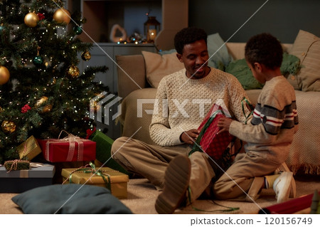 Full length portrait of happy African American family of caring father and son opening Christmas presents together sitting on floor Full length portrait of happy African American family of caring father and son opening Christmas presents together sitting on floor 120156749