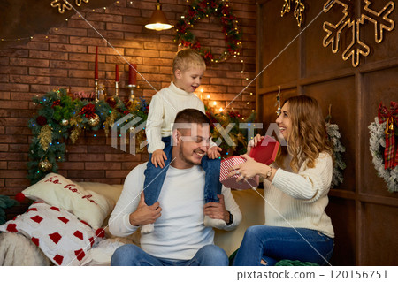 Happy family opening presents next to Christmas tree and smiling cheerfully on Christmas Eve in a Happy family opening presents next to Christmas tree and smiling cheerfully on Christmas Eve in a 120156751