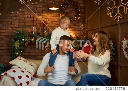 Happy family opening presents next to Christmas tree and smiling cheerfully on Christmas Eve in a 120156756