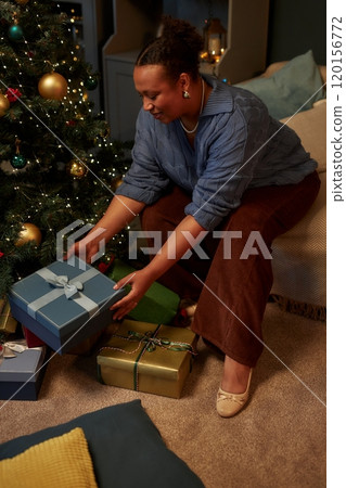 Vertical side view portrait of smiling adult African American woman opening Christmas presents in elegant home setting 120156772