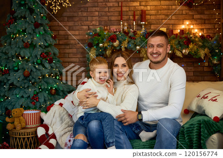 Happy family hugging warmly next to Christmas tree and smiling cheerfully on Christmas Eve in a Happy family hugging warmly next to Christmas tree and smiling cheerfully on Christmas Eve in a 120156774