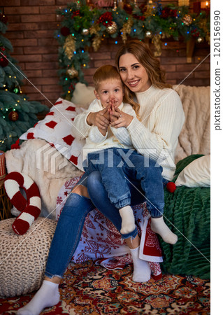 Happy mom and little child sitting on sofa near Xmas tree, together at home while spending leisure Happy mom and little child sitting on sofa near Xmas tree, together at home while spending leisure 120156990