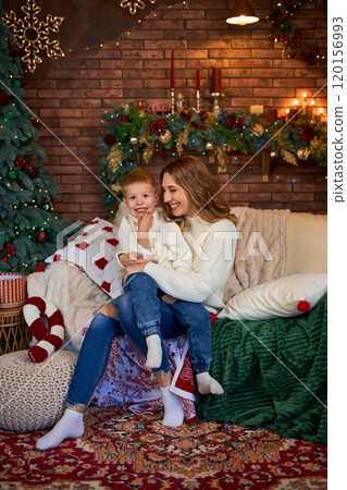 Happy mom and little child sitting on sofa near Xmas tree, together at home while spending leisure Happy mom and little child sitting on sofa near Xmas tree, together at home while spending leisure 120156993