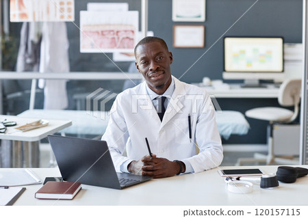 Front view portrait of friendly African American doctor smiling at camera while sitting at workplace in professional clinic setting copy space Front view portrait of friendly African American doctor smiling at camera while sitting at workplace in professional clinic setting copy space 120157115
