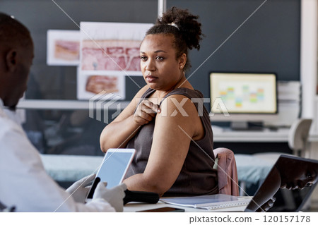 Portrait of adult African American woman as patient in dermatology clinic consulting with doctor and pointing at shoulder copy space Portrait of adult African American woman as patient in dermatology clinic consulting with doctor and pointing at shoulder copy space 120157178